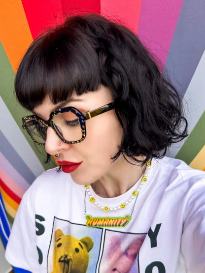 Artist, Stoofy with short dark hair and glasses against a colourful geometric background, wearing the ‘Humanity’ pendant necklace. Design and copyright by Stoofy.