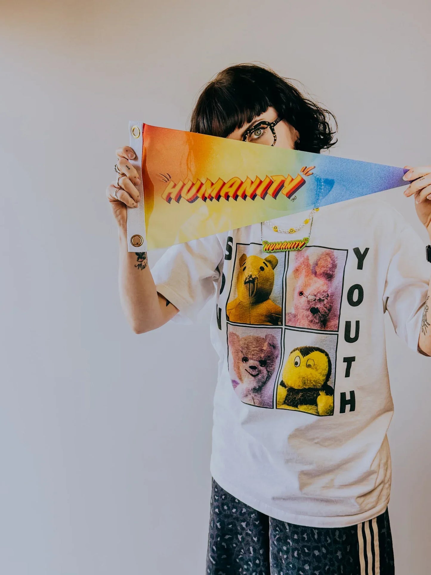 Artist, Stoofy holding  rainbow coloured ‘Humanity’ pennant flag, wearing a white t-shirt with graphic design. Design and copyright by Stoofy. 