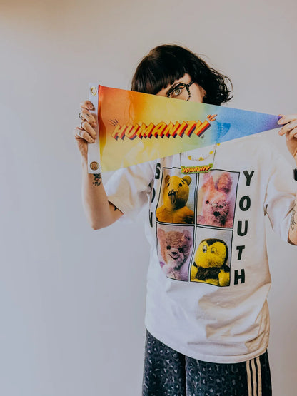 Artist, Stoofy holding  rainbow coloured ‘Humanity’ pennant flag, wearing a white t-shirt with graphic design. Design and copyright by Stoofy. 