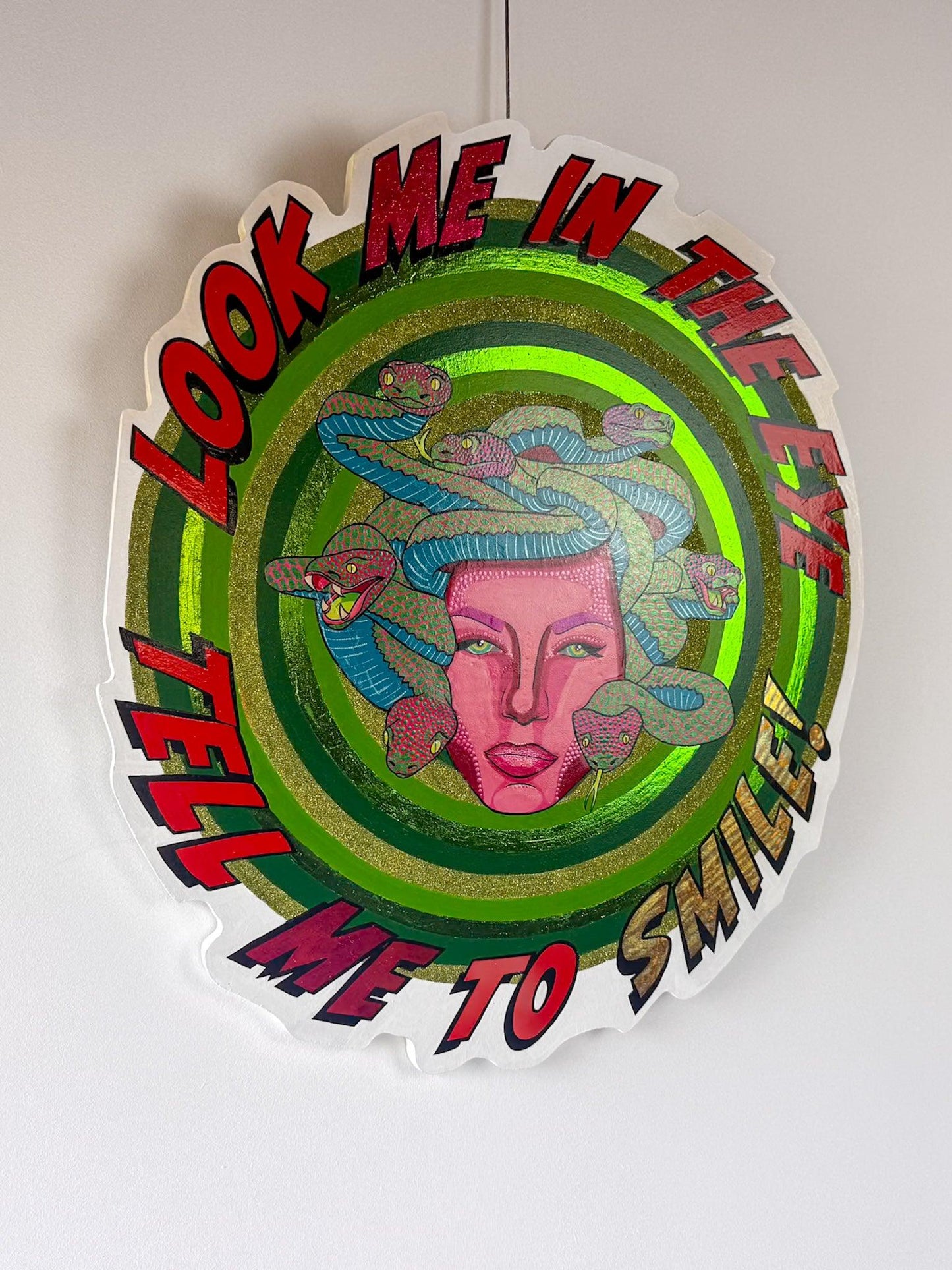 Angled view of Look Me in the Eye — Tell Me to Smile! by Stoofy, showing the metallic foil ring catching the light. The brightest green-gold foil band glows against the surrounding hand-painted concentric rings. Medusa at centre with magenta pink face, Ben-Day dots and multiple snakes in blue and pink. Lettering around the edge in red and gold reads "Look Me in the Eye — Tell Me to Smile!" Original mixed media on hand-cut wood panel. © Stoofy