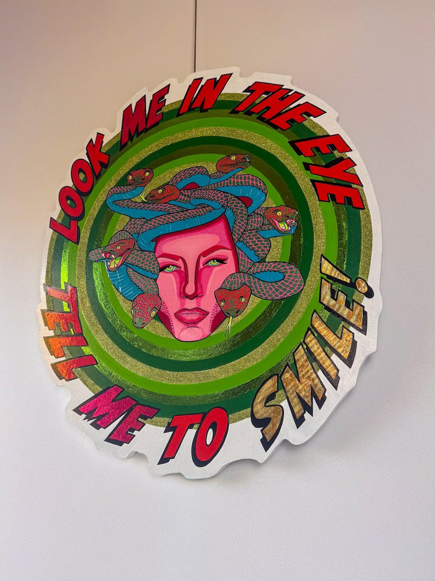 Front-on full view of Look Me in the Eye — Tell Me to Smile! by Stoofy. Circular hand-cut shaped wood panel showing Medusa with a calm magenta pink face and green-gold eyes, her head entwined with blue and red-pink snakes. Concentric painted green rings with glitter and foil surround her. Bold red and gold lettering arcs around the scalloped white edge. Original mixed media artwork. One of one. © Stoofy