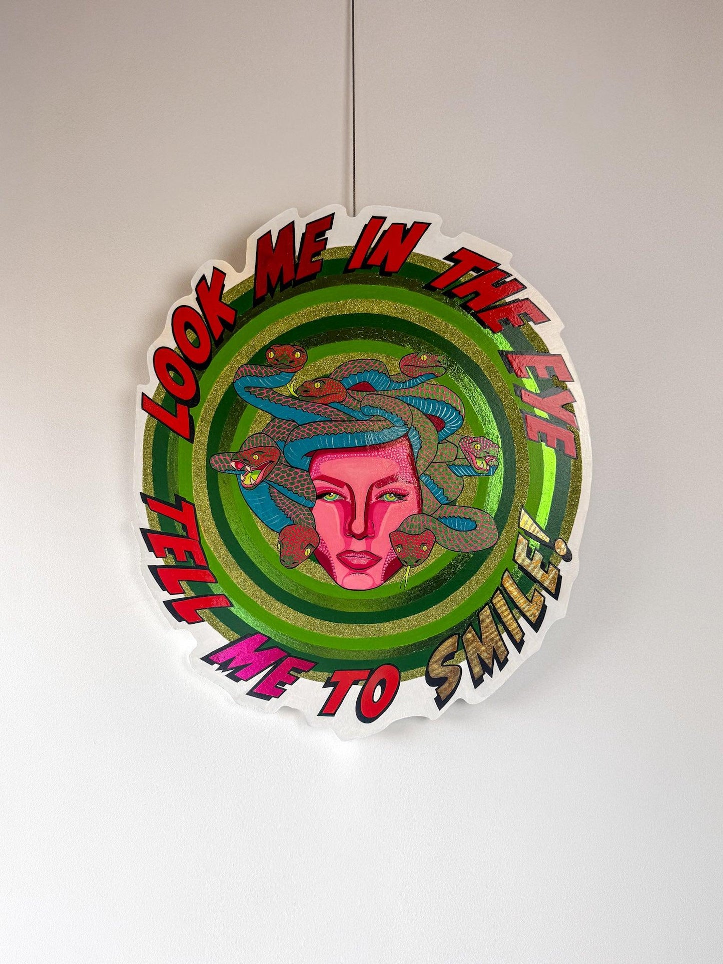 Full view of Look Me in the Eye — Tell Me to Smile! by Stoofy, hanging against a white wall. A circular hand-cut wood panel painted with Medusa at centre — a magenta pink face with Ben-Day dot detailing, surrounded by blue, pink and red snakes with individually painted scales. Concentric rings of hand-painted green with gold-green glitter and metallic foil radiate outward. Bold comic-style lettering circles the edge reading "Look Me in the Eye — Tell Me to Smile!" Mixed media original. One of one. © Stoofy
