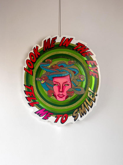 Full view of Look Me in the Eye — Tell Me to Smile! by Stoofy, hanging against a white wall. A circular hand-cut wood panel painted with Medusa at centre — a magenta pink face with Ben-Day dot detailing, surrounded by blue, pink and red snakes with individually painted scales. Concentric rings of hand-painted green with gold-green glitter and metallic foil radiate outward. Bold comic-style lettering circles the edge reading "Look Me in the Eye — Tell Me to Smile!" Mixed media original. One of one. © Stoofy
