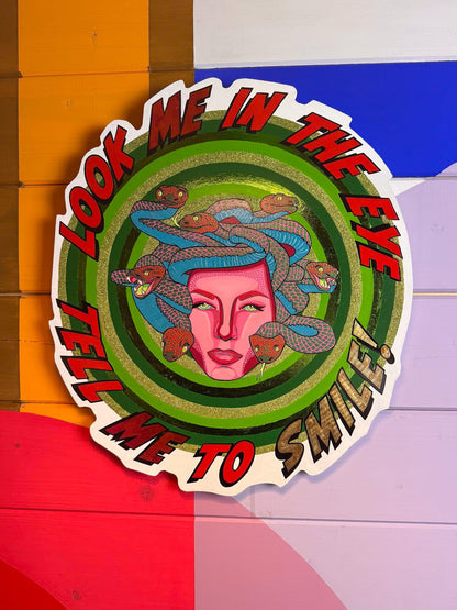 Look Me in the Eye Tell Me to Smile! by © Stoofy mounted against a bold colour-block painted wood panel wall in orange, blue, red, lavender and white geometric sections. The circular hand-cut wood panel shows Medusa at centre — a magenta pink face with Ben-Day dot detailing and calm green-gold eyes, surrounded by blue and red-pink snakes with individually painted scales. Concentric hand-painted green rings with gold-green glitter and metallic foil radiate outward. Bold red and gold comic-style lettering arc