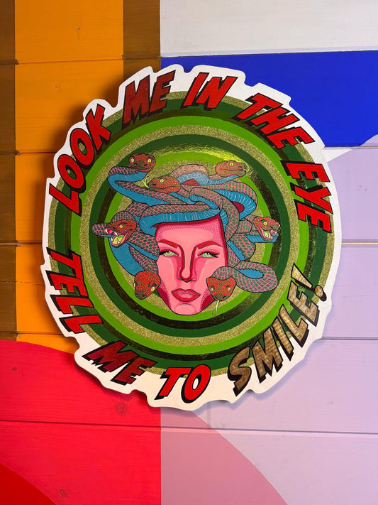 Look Me in the Eye Tell Me to Smile! by © Stoofy mounted against a bold colour-block painted wood panel wall in orange, blue, red, lavender and white geometric sections. The circular hand-cut wood panel shows Medusa at centre — a magenta pink face with Ben-Day dot detailing and calm green-gold eyes, surrounded by blue and red-pink snakes with individually painted scales. Concentric hand-painted green rings with gold-green glitter and metallic foil radiate outward. Bold red and gold comic-style lettering arc