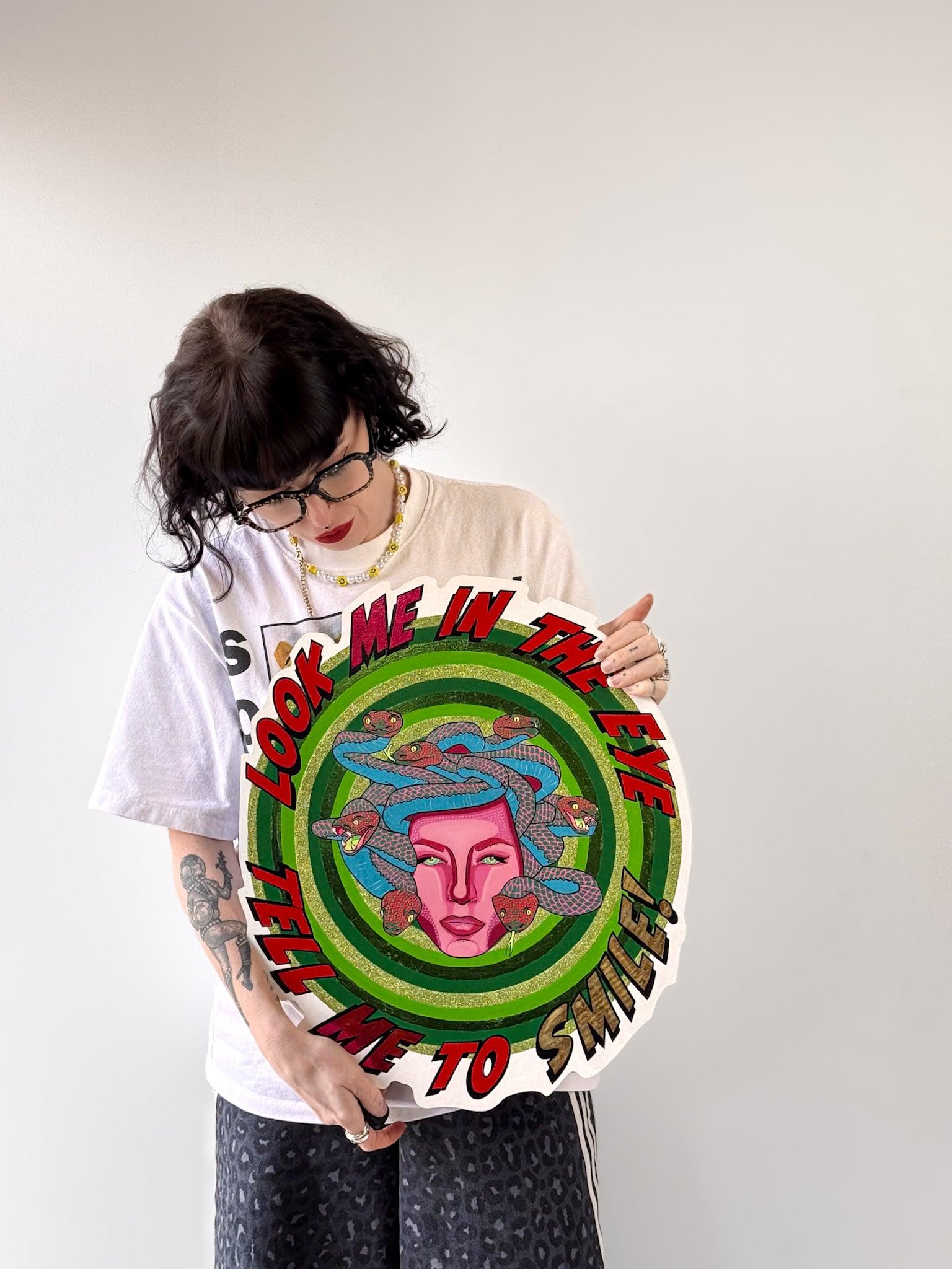 Artist and founder of Stoofy, holding Look Me in the Eye — Tell Me to Smile! against a pale background. She wears glasses, a white t-shirt, dark trousers and a beaded necklace, with tattoos visible on her left arm. She looks down at the piece. The circular hand-cut wood panel shows Medusa at centre with a magenta pink face and blue and red-pink snakes, surrounded by concentric green rings with gold-green glitter. Original mixed media artwork. Made in the UK. © Stoofy
