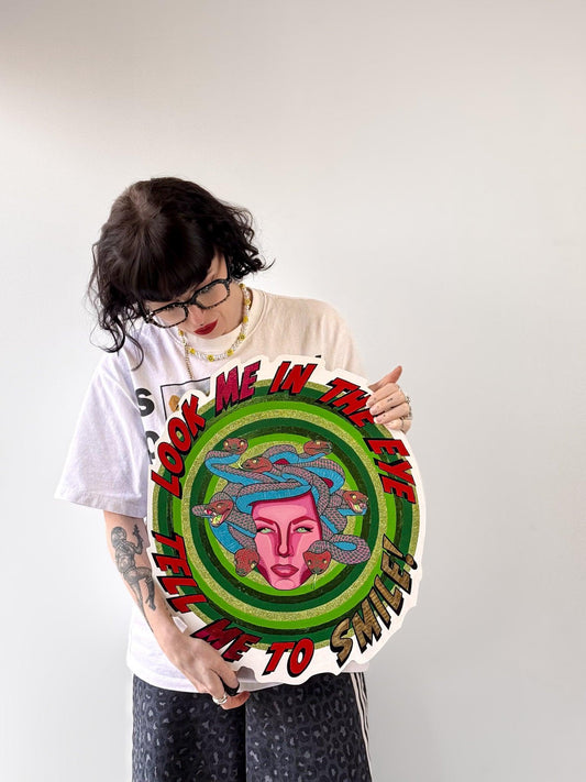 Artist and founder of Stoofy, holding Look Me in the Eye — Tell Me to Smile! against a pale background. She wears glasses, a white t-shirt, dark trousers and a beaded necklace, with tattoos visible on her left arm. She looks down at the piece. The circular hand-cut wood panel shows Medusa at centre with a magenta pink face and blue and red-pink snakes, surrounded by concentric green rings with gold-green glitter. Original mixed media artwork. Made in the UK. © Stoofy
